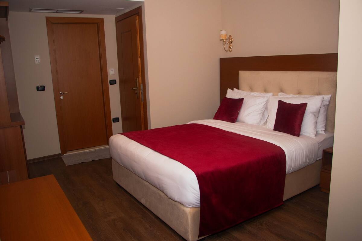 A hotel room with a bed, a wooden nightstand, a wooden desk, and a door.