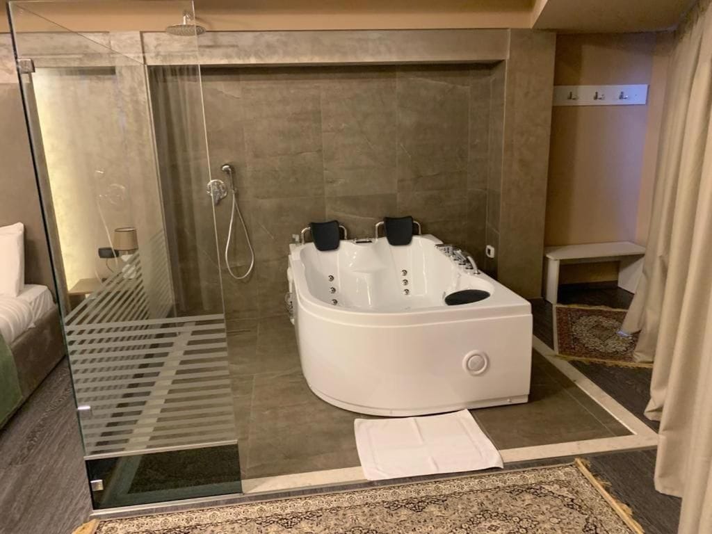 Un baño moderno con ducha envuelta en vidrio, una bañera blanca con múltiples chorros y una mesita de noche pequeña.