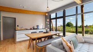A modern kitchen with a wooden table and chairs, a sofa with cushions, and large windows offering a view of the outdoors.