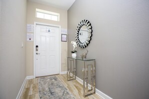 A hallway with a white door, a glass console table with a decorative mirror, and a patterned rug.