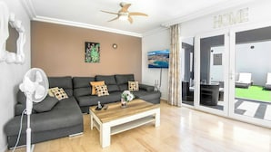 Ein modernes Wohnzimmer mit einem grauen Sofa, einem Holztisch, einem Standventilator und einem an der Wand befestigten Fernseher.
