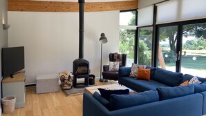 A modern living room with a wood-burning stove, a blue sofa, a television, and large windows offering a view of the outdoors.