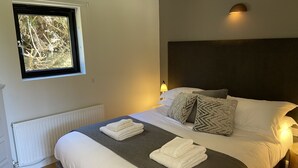 A neatly made bed with white and grey linens, a window with a view of trees, and a wall-mounted lamp.