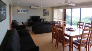 Un vaste espace de vie comprenant un canapé noir, une table à manger avec des chaises en bois, un téléviseur et un ventilateur de plafond.