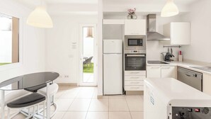 Een moderne keuken met witte kasten, een ingebouwde oven, een magnetron en een vaatwasser. Er is een eettafel met stoelen en hanglampen erboven.