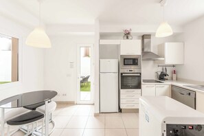 Una cocina moderna con armarios blancos, horno integrado, microondas y lavavajillas. Hay una mesa de comedor con sillas y lámparas colgantes arriba.