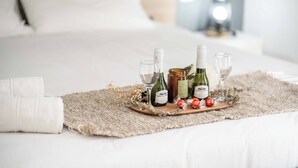 Una cama bien hecha con una bandeja de madera que sostiene botellas de vino y copas, una toalla blanca enrollada y detalles decorativos.