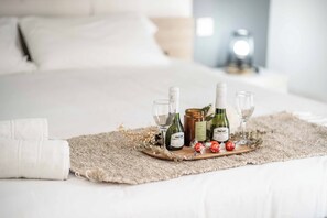 Un lit bien fait, sur lequel repose un plateau en bois contenant des bouteilles de vin et des verres, une serviette blanche roulée et quelques éléments décoratifs.
