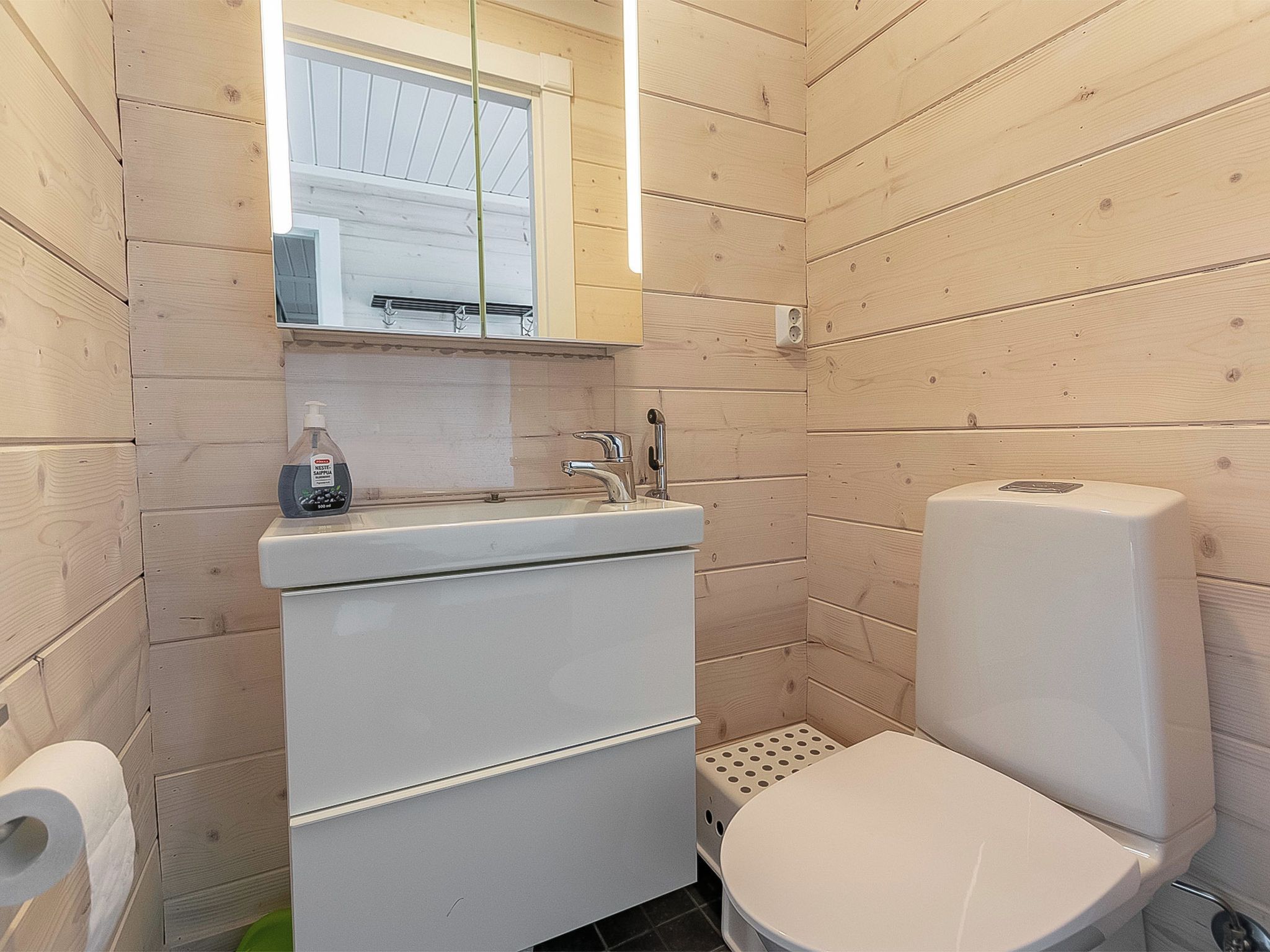 Bany compacte amb vàter blanc, un lavabo amb mirall i una ampolla de sabó de mans a la superfície.