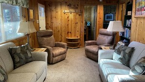 A cozy cabin interior with wooden walls, two sofas, two armchairs, a small table, a lamp, and a bookshelf.