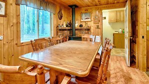 A wooden dining room with a large table and chairs, a fireplace, and a kitchen area visible through an open door.