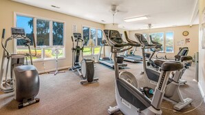 Ein Fitnessstudio mit unterschiedlichen Trainingsgeräten, großen Fenstern und einem Deckenventilator.