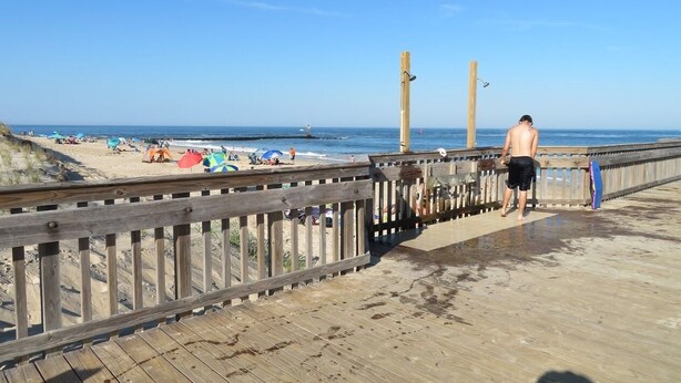 Ein hölzerner Steg führt zu einem Strand mit Sonnenschirmen und Menschen.