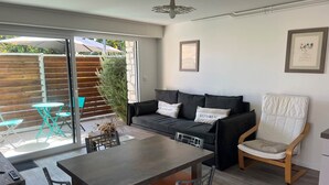 A modern living room with a sofa, a dining table, and a chair. There is a sliding glass door leading to an outdoor area with a table and chairs.