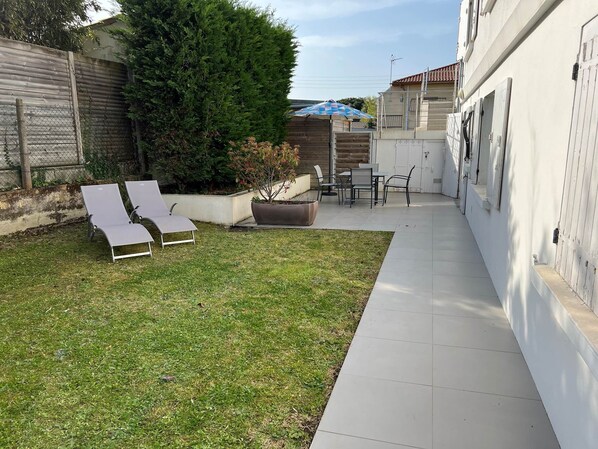 A patio area with two lounge chairs, a dining table with chairs, a potted plant, and a large tree. - Nice rent with garden near the beach and city center (ST GEORGES DE DIDONNE)