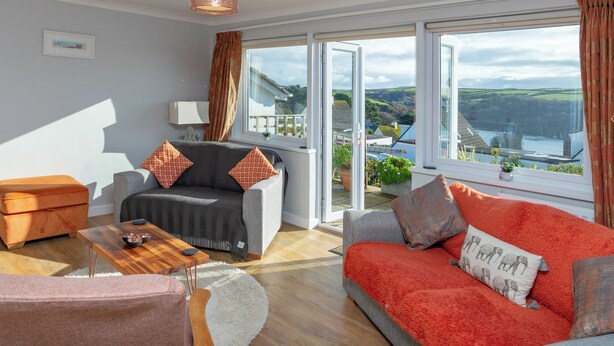 A modern living room with a grey sofa, orange cushions, a red sofa, and a wooden coffee table. There is a large window with a view of the countryside. - 2 bedroom accommodation in Fowey (Fowey)