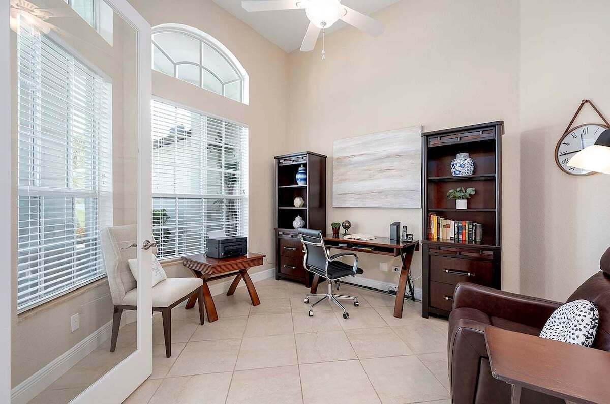 A home office with a desk, chair, bookshelf, and a large window with blinds.