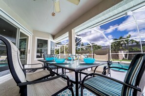 Een terras buiten met een glazen tafel en stoelen, een zwembad en uitzicht op palmbomen en een bewolkte lucht.
