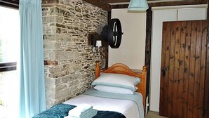 A bedroom with a stone wall, wooden ceiling beams, a bed with white and blue bedding, a hanging lamp, and a wooden door. - 2 bedroom accommodation in Fowey (Fowey)