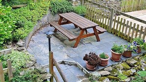A wooden picnic table with benches, surrounded by a stone wall and a wooden fence. There are potted plants and a stone pathway. - 2 bedroom accommodation in Fowey (Fowey)