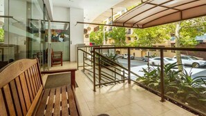 Un balcon avec un banc en bois, des portes vitrées et une vue sur une rue animée, avec des voitures et des immeubles.