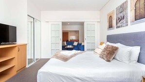 Une chambre d’hôtel moderne avec un grand lit, un téléviseur posé sur un meuble en bois, une armoire et un coin salon avec un canapé bleu et une table.