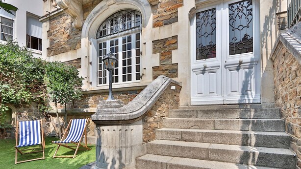 Un escalier en pierre mène à une porte blanche ornée d’un portail en fer forgé, le tout étant entouré d’un mur de pierre et d’un petit jardin où se trouvent deux chaises de jardin à rayures.