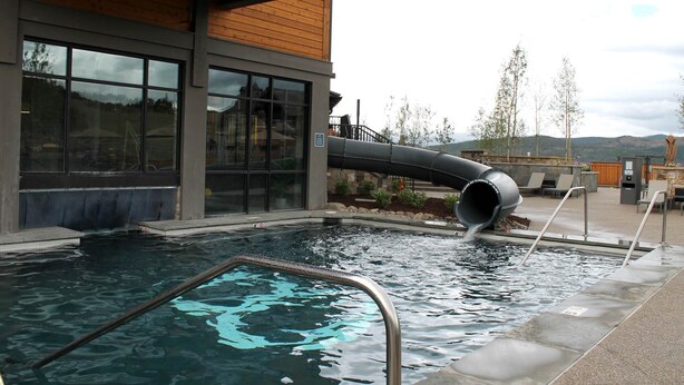 A pool with a slide, surrounded by a building with large windows and a view of hills. - The Premier Ski-in Ski-out Address in all of Colorado - Unit LIcense 81047001. (Breckenridge)