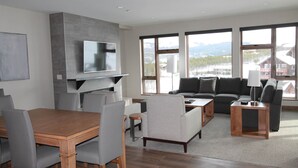 A modern living room with a wooden dining table, grey upholstered chairs, a grey sofa, and a TV mounted on a grey wall. - The Premier Ski-in Ski-out Address in all of Colorado - Unit LIcense 81047001. (Breckenridge)
