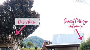 A gravel driveway leading to a house with a stone wall and a large tree. An arrow points to the house labeled "Our House," and another points to a building with a metal roof labeled "Sunset Cottage entrance." - Sunset Cottage with Beautiful Blue Ridge Mountain Views (Clyde)