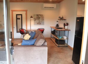 A compact living space with a sofa, chairs, a small table, a kitchen area with a refrigerator, and a wall-mounted air conditioner.