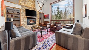 Una sala de estar moderna con chimenea de piedra, sofás de cuero, televisor de pantalla plana y un ventanal con vistas al bosque.
