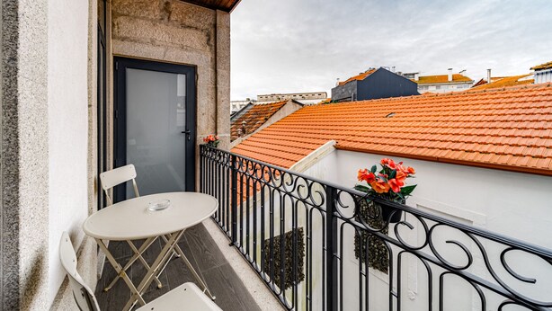 Un balcón con mesa y sillas, una puerta y vistas a edificios vecinos con tejados anaranjados.