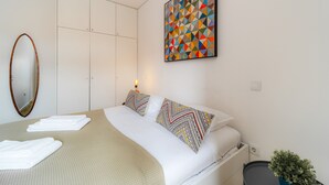 Una cama bien hecha con ropa blanca y almohadas estampadas, un cuadro abstracto grande en la pared y una pequeña planta en maceta en el suelo.