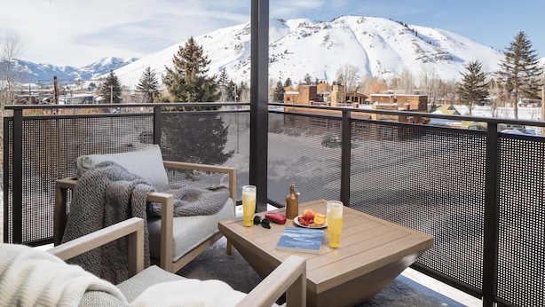 Ein Balkon mit Blick auf schneebedeckte Berge, ein Holztisch mit Getränken und einer Speisekarte sowie ein Sessel mit Decke.