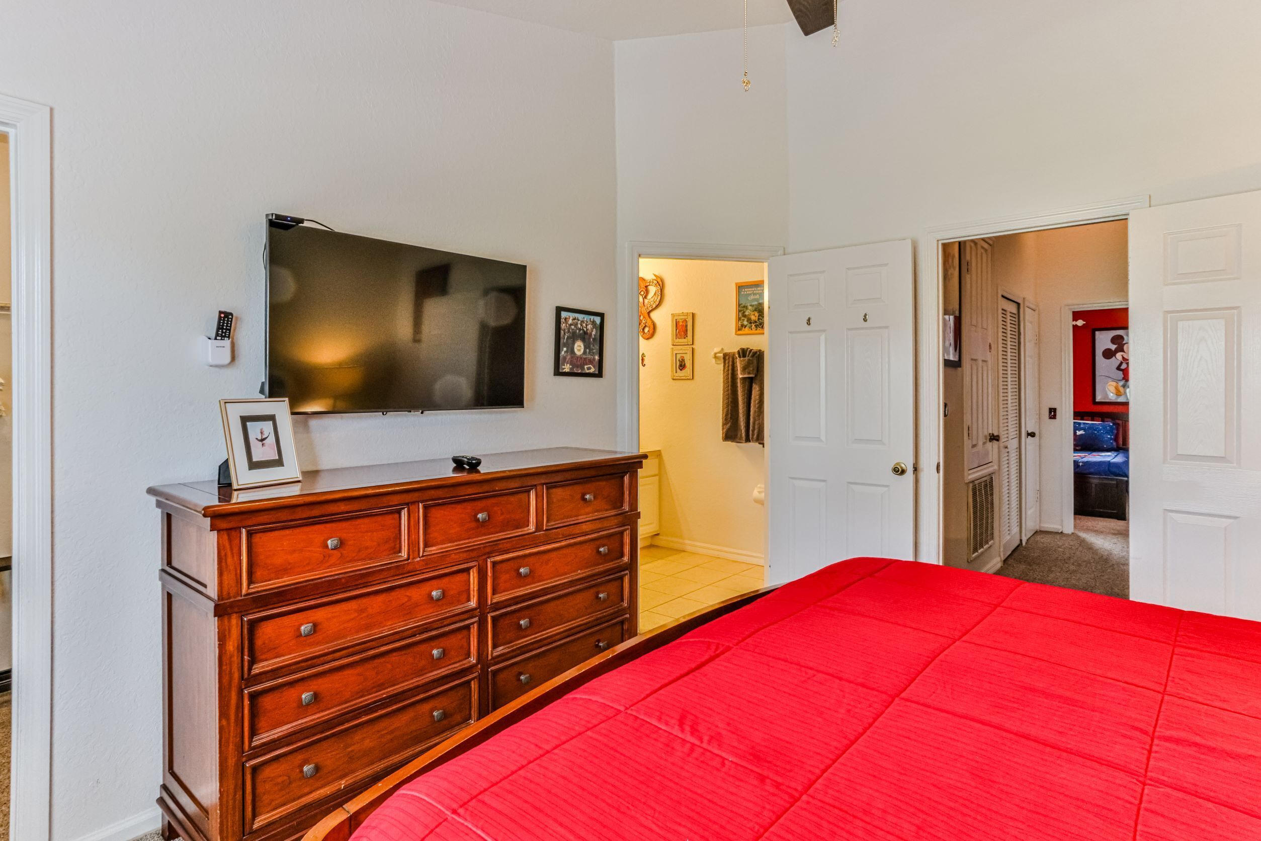 Un dormitorio con edredón rojo, cómoda de madera, televisor de pantalla plana y pasillo que conduce a otra habitación.