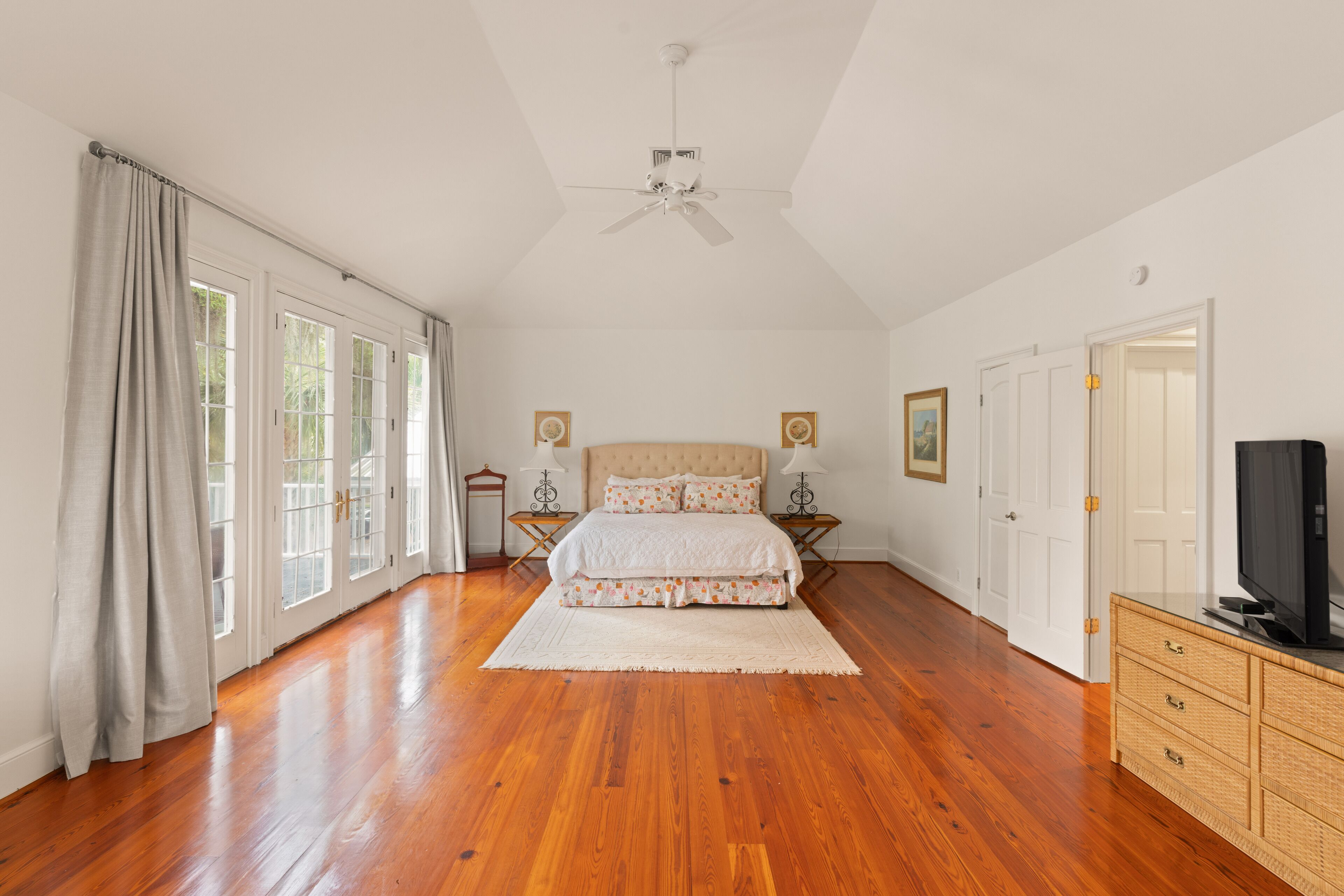 Une chambre spacieuse avec un plancher en bois, un grand lit, un ventilateur de plafond et un téléviseur posé sur une commode.