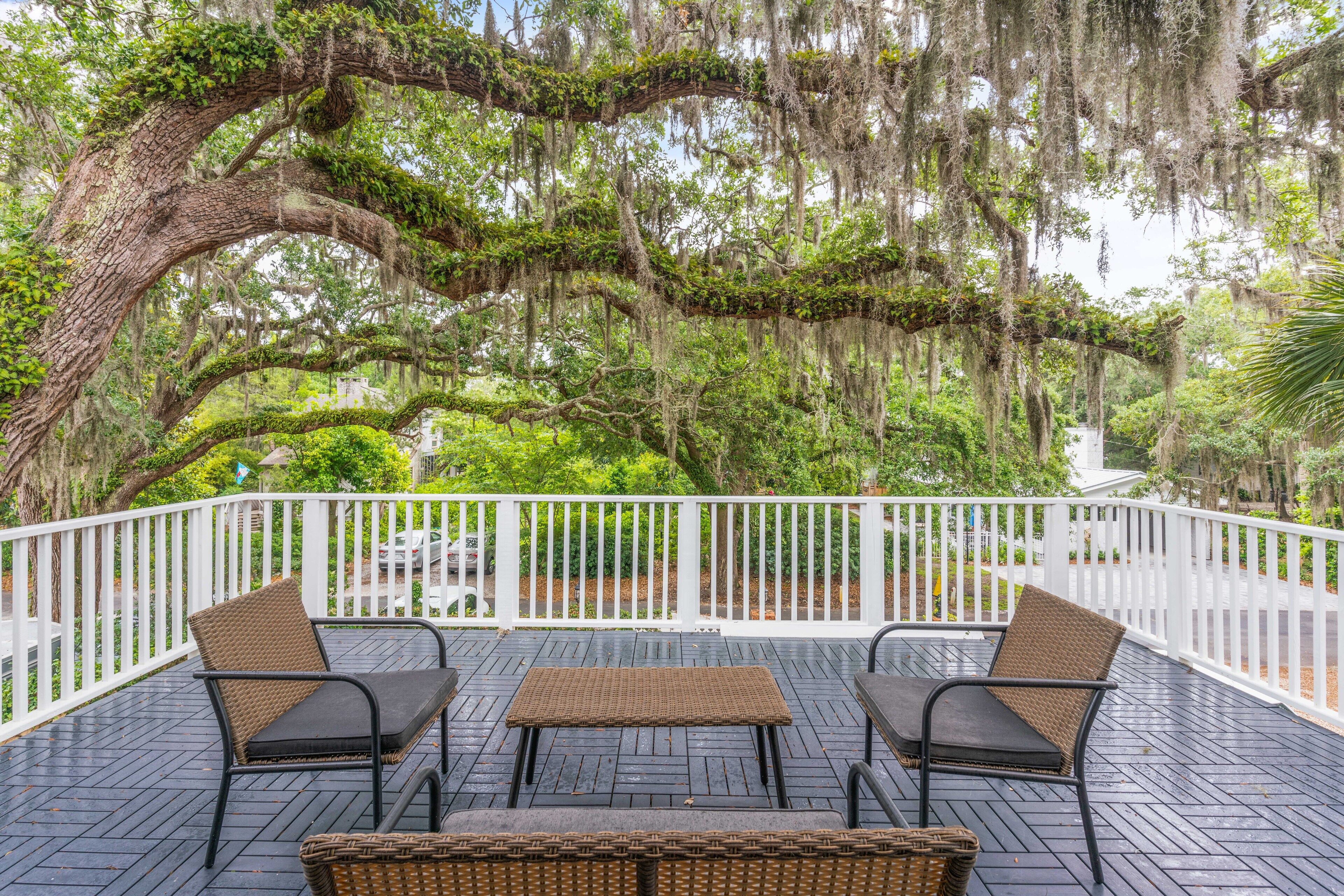 A covered outdoor deck with wicker furniture, surrounded by a white fence and large trees with hanging moss.