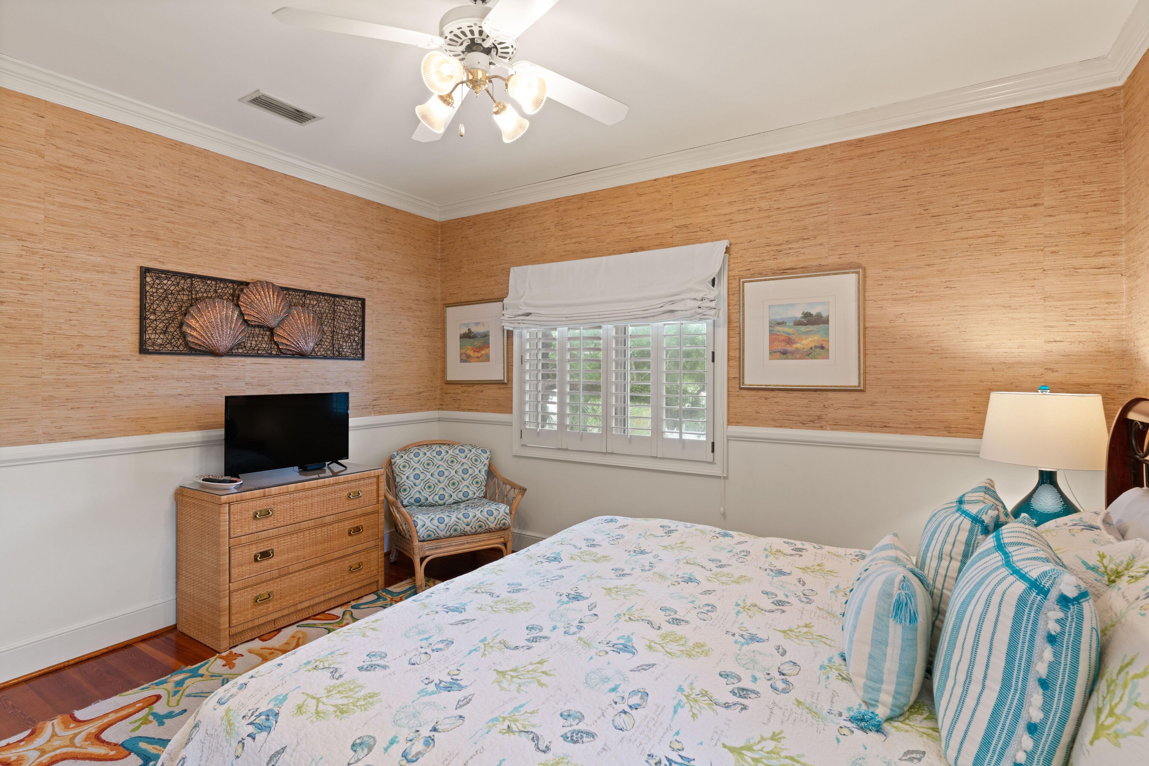 Une chambre à coucher avec un lit, un téléviseur posé sur une commode, une chaise, une fenêtre avec des volets et des œuvres d’art accrochées aux murs.