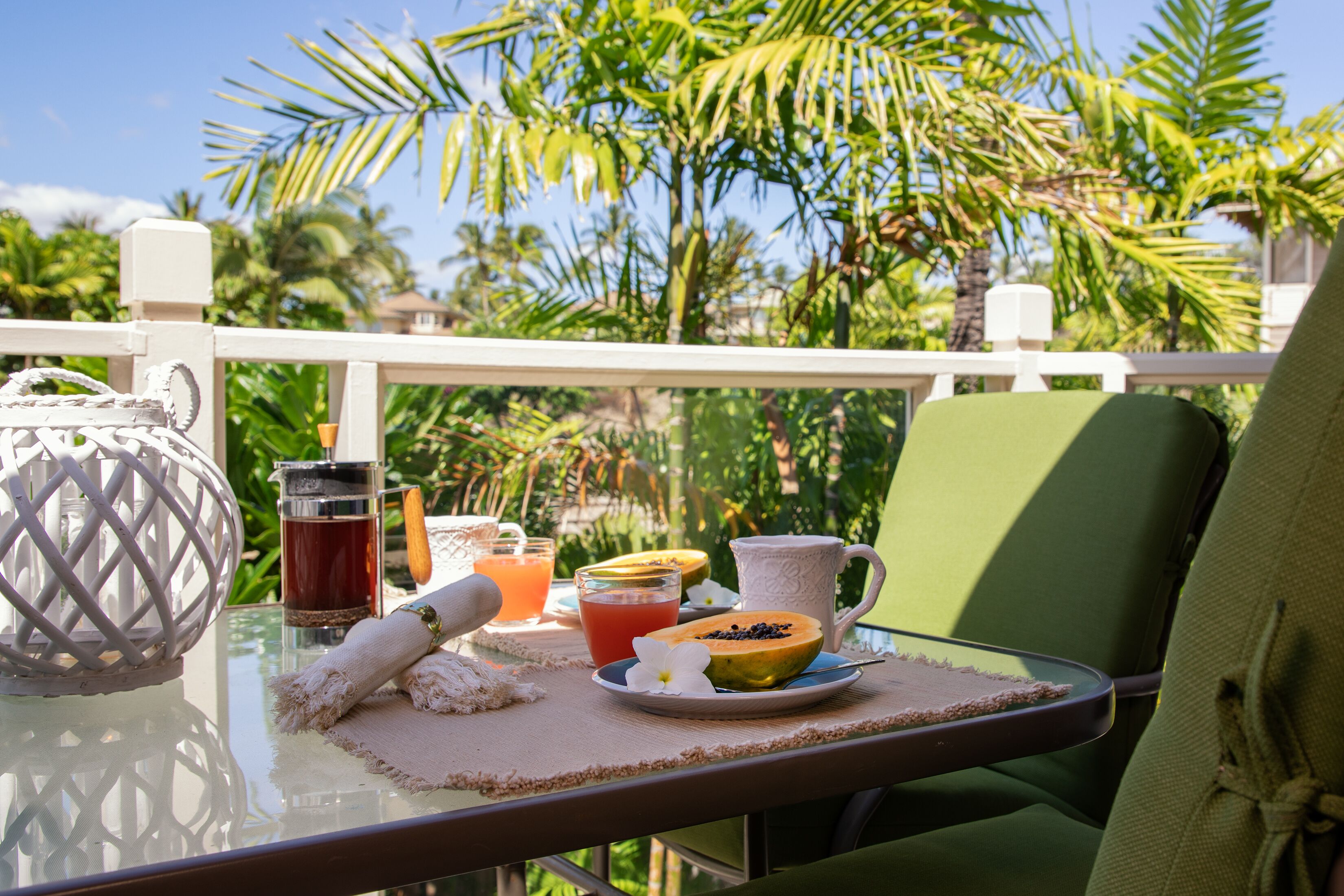 Une table avec un verre de thé glacé, un saladier de fruits et une tasse blanche sur un balcon d’où l’on peut admirer des palmiers.