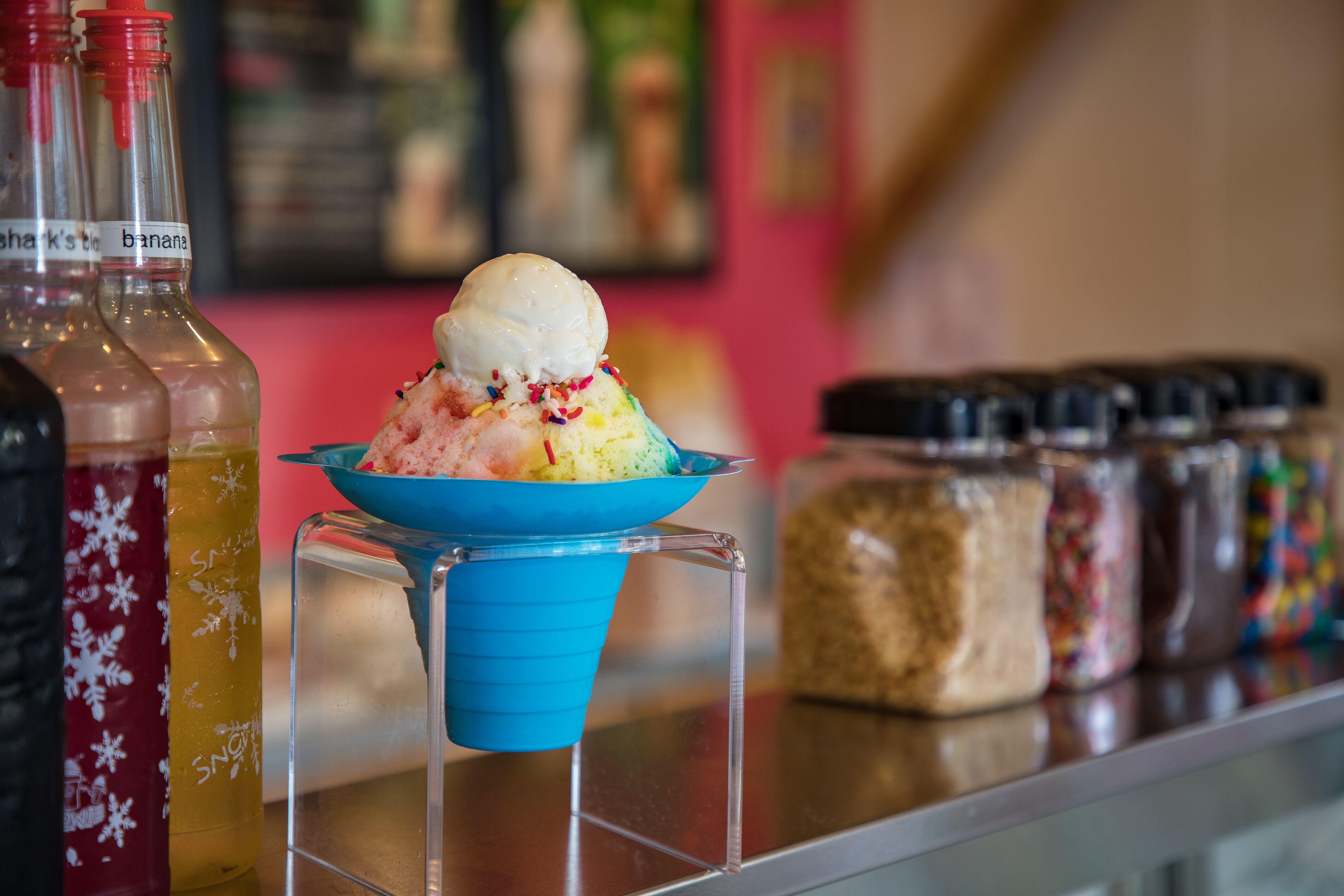 Une coupe glacée colorée, surmontée d’une boule de glace à la vanille, servie dans un bol bleu posé sur un présentoir transparent.