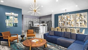 A modern living room with a blue sofa, two armchairs, a coffee table, and a dining area with a large painting on the wall.