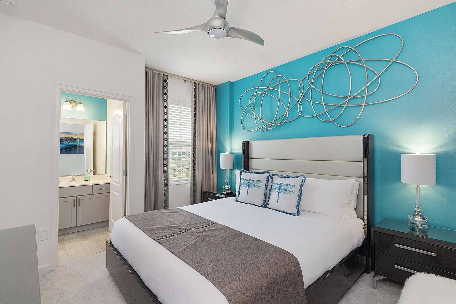 A modern hotel room with a large bed, a ceiling fan, a bathroom with a vanity, and a view of the city through the window.