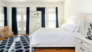 Un dormitorio con una cama, ventilador de techo, mesita de noche, lámpara, una silla de mimbre y un tapete estampado.