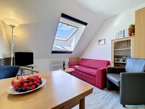 Un appartement moderne de style « loft » avec un plafond en pente, un canapé rouge, une table à manger avec des fruits, un téléviseur et une lucarne.
