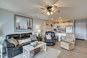A living room with a black leather sofa, a white coffee table, and a beige armchair. There is a ceiling fan, a painting of sailboats, and a kitchen in the background.
