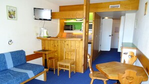A compact room with a blue sofa, wooden dining table, and chairs, a small kitchen area with a microwave, and a television mounted on the wall.