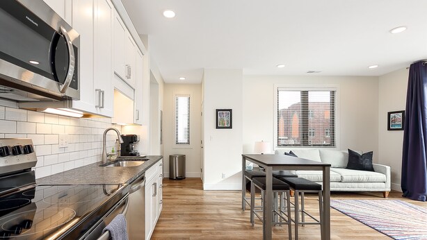 A modern kitchen with stainless steel appliances, a dining area with a table and chairs, and a living room with a sofa and a window. - Modern Downtown Masterpiece (Traverse City)