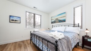 A bedroom with a bed, two windows with blinds, a nightstand with a lamp, and two framed pictures on the wall. - Modern Downtown Masterpiece (Traverse City)