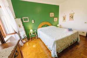 A room with a green wall, wooden floor, a bed with a light blue bedspread, a bedside table with a lamp, and framed pictures on the wall.